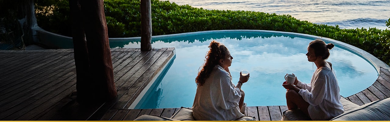 Two women seated by a pool overlooking lush greenery and the ocean, enjoying drinks at sunset.