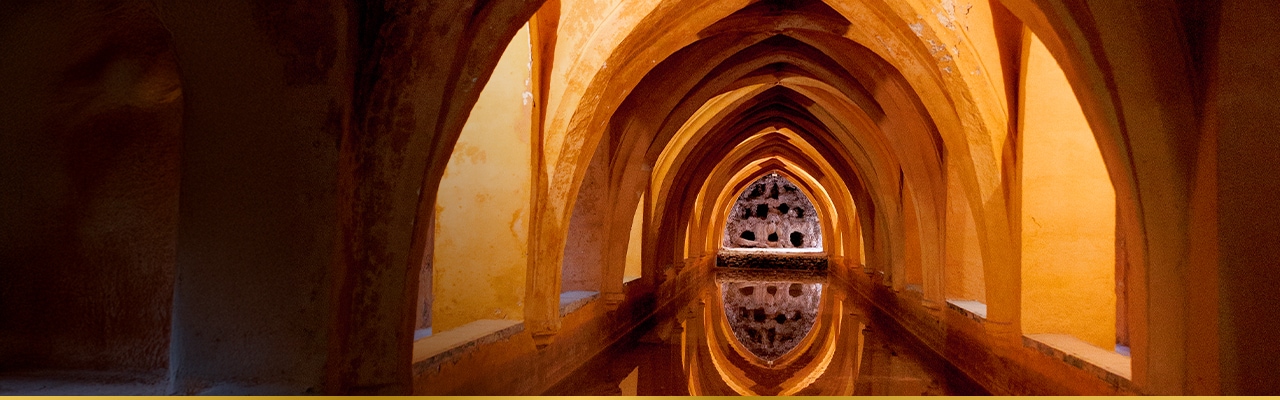 Underground arched corridor with golden lighting reflecting on a still pool of water.